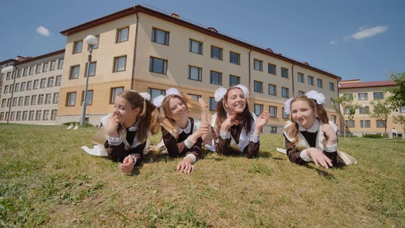 Four Schoolgirl Graduates are Lying on the Grass Against the Backdrop of Their School