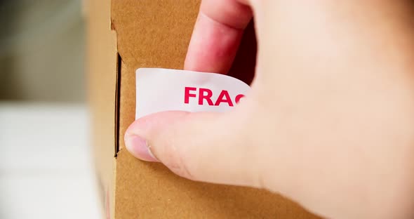 Hands applying FRAGILE Sticker label on a cardboard product box with barcode this side up.