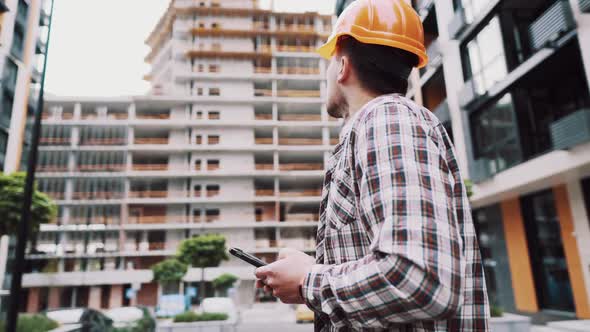 Caucasian Male Construction Worker in Protective Helmet Use Smartphone Near Building That Is Being