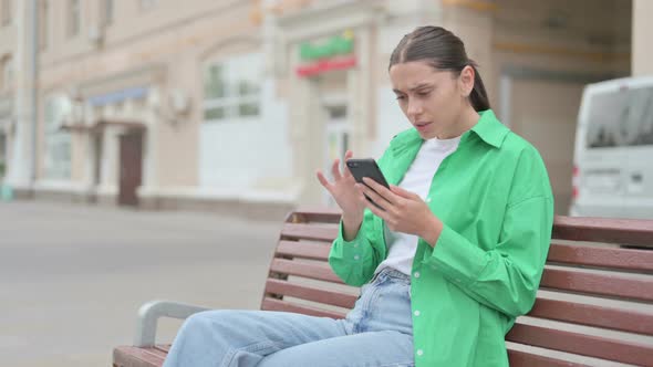 Hispanic Woman Reacting to Loss on Smartphone While Sitting Outdoor on Bench
