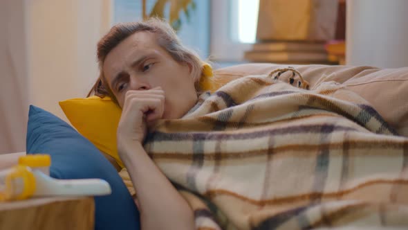 Sick Young Man Lying on Couch Covered with Blanket and Coughing