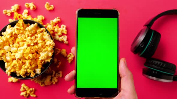 Man's hand holding smart phone with green touch screen and popcorn in a bowl on red background.