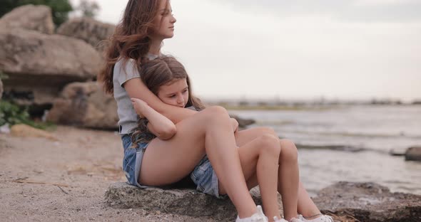 Two Cute Sisters Sitting at the Beach of the Sea and Relaxing
