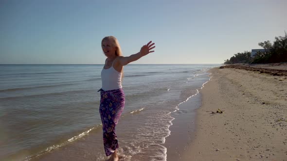 Slow motion of joyous mature woman backlit, splashing in the water on a beach at sunrise or sunset.