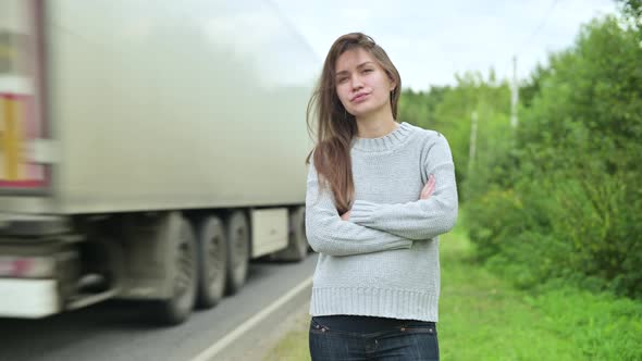 A large truck drives quickly past a young woman who is waiting for transport