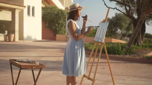 Slim Caucasian Woman in Dress and Straw Hat Painting on Canvas Drinking Red Wine From Glass Outdoors