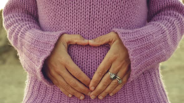 Woman hands do hearth sign on belly against violet sweater. Romantic and newborn life pregnant