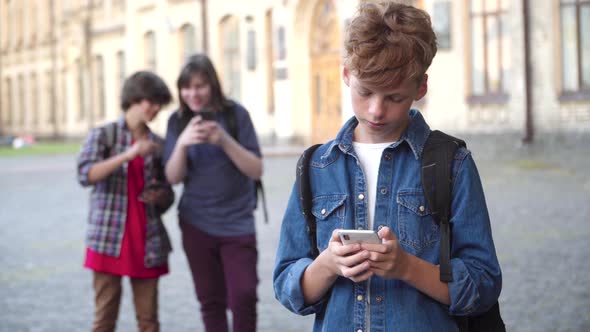 Sad Redhead Caucasian Schoolboy Using Smartphone, Sighing. and Looking Back at Classmates Teasing