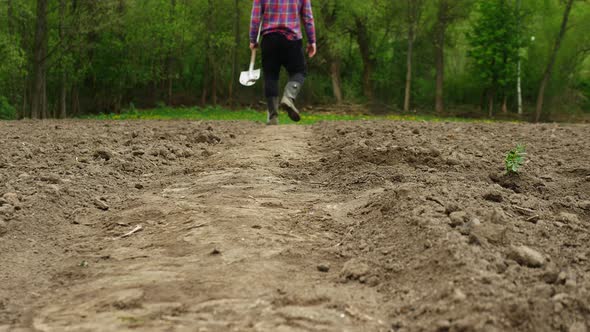 Farmer With A Shovel In His Hand Walks On The Black Soil In The Garden. The Farmer Takes Black Soil