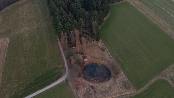 A small, round pond between green fields on the edge of a coniferous forest in Germany. Aerial fly o