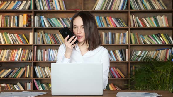 Brunette Woman Talks on Phone Near Laptop at Table in Room