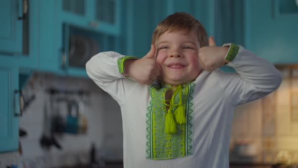 Blonde boy in embroidered national clothes smiling and showing thumbs up at home