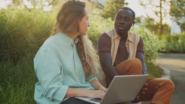 Multiethnic Man and Woman Sitting with Laptop on Street and Speaking