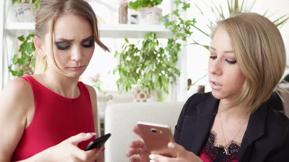 Two Young Girlfriends Using Smartphone and Talking in Cafe and Drinking Coffee