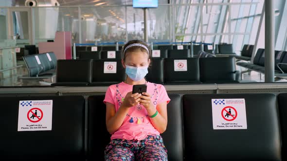 Teenage Girl, in Protective Mask, Uses Mobile. She Sitting in an Empty Airport Lounge, Waiting 