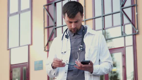 Young Doctor in a White Lab Coat Wears Eyeglasses and Surfs on His Phone Messages As He Leaves the