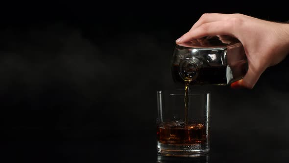 Barman Pour Whiskey Cognac Brandy From Bottle Into Drinking Glass with Ice Cubes on Dark Background