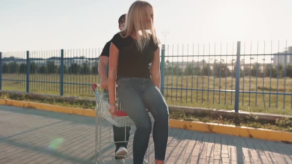 The Guy Rolls Attractive Young Woman in the Trolley on Parking Lot of the Supermarket at Sunset