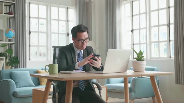 Asian Businessman Using Mobile Phone While Using The Computer For Working At Home