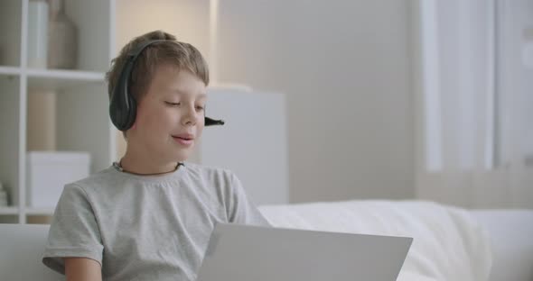 Cute Little Boy Is Chatting with Friend By Internet Using Laptop and Wireless Headphones From Home