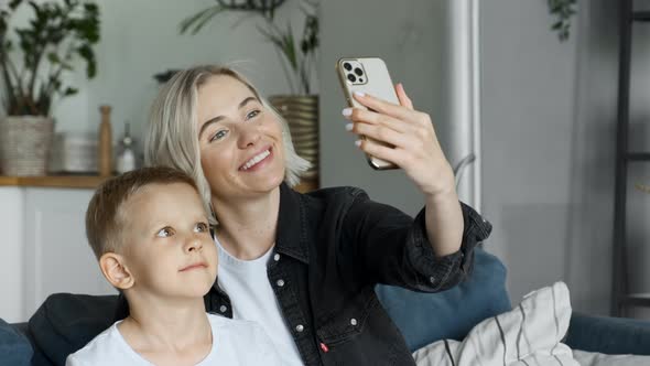 Mother taking selfie with boy using mobile phone. Woman and child sitting on the sofa and make video