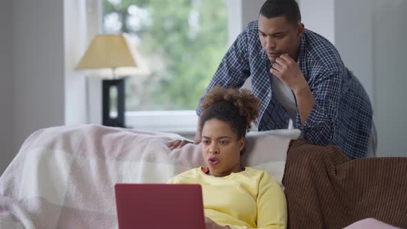 Focused African American Woman Messaging Online on Laptop As Worried Man Supporting Wife Talking and