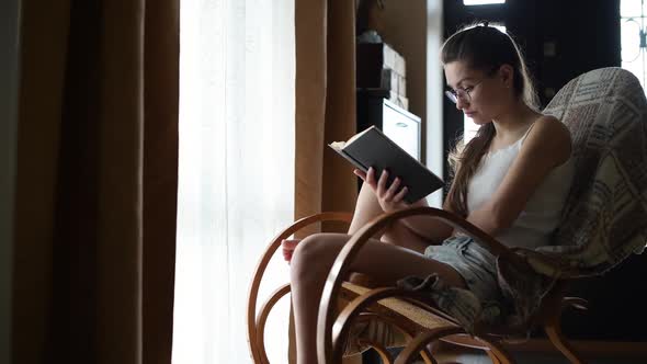 woman reads a book sitting in a rocking chair in front of the window