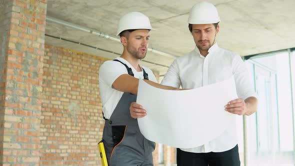 Two Young Engineers in Safety Helmets with a Design Plan Standing on the Construction Site