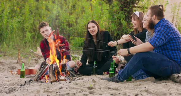 Group of Young Friends Sitting By the Fire at Autumn Beach Grilling Sausages and Drinking Beer