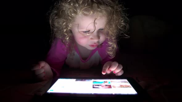 Close up of adorable curly-haired girl of four years old uses a tablet at night in bed.