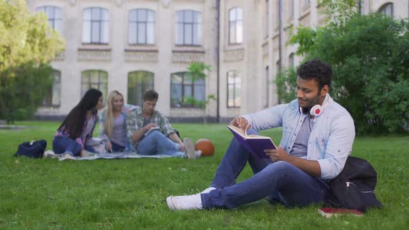 Happy Biracial Male Student Sitting on Grass and Reading Interesting Book