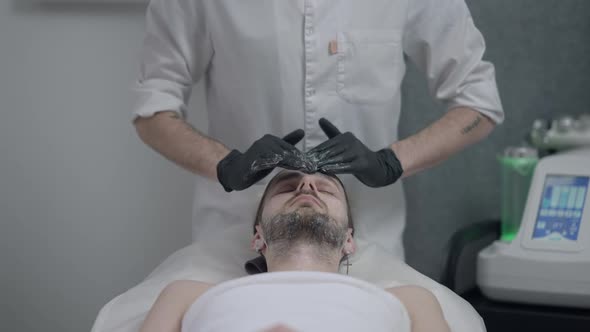 Young Handsome Caucasian Man Lying in Beauty Salon with Closed Eyes As Male Hands in Black Gloves