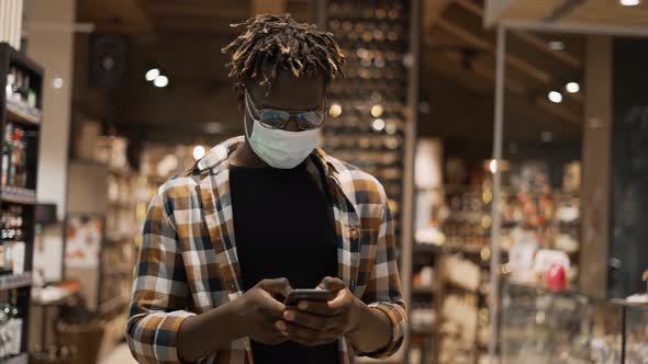 African American Man in Mask Busy with Smartphone Walks at the Store