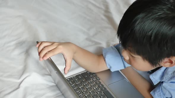 Cute Asian Boy Lying In Bed And Using Laptop On White Bed