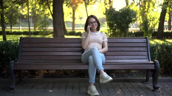 A Young Woman in Glasses Sits on a Bench Speaks on the Phone. In the Background Nature