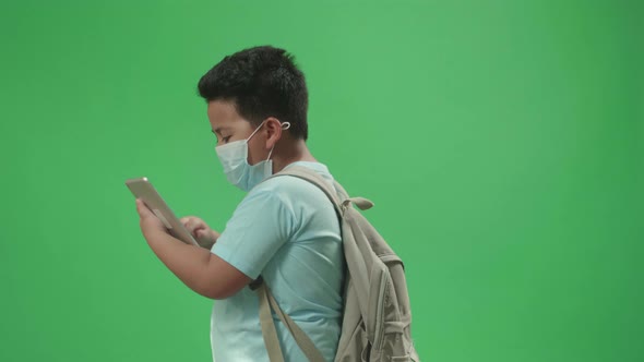 Side View Of Asian Boy Student Wearing A Mask And Use Tablet While Walking On Green Screen