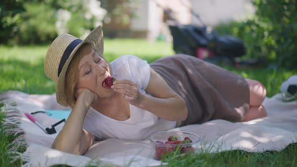 Relaxed Senior Woman Tasting Delicious Strawberry in Slow Motion Lying on Blanket on Sunny Backyard