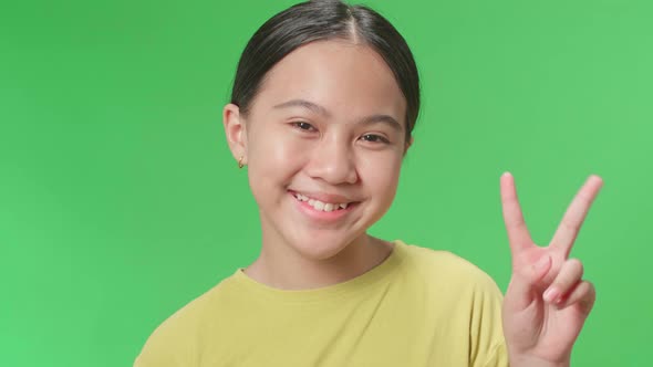 A Smiling Young Asian Kid Girl Showing Gesture Peace While Standing On Green Screen In The Studio alt