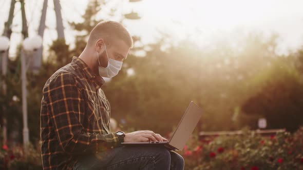 Masked Man is Typing on Laptop While Sitting Park Bench Turns His Head Takes Off Mask and Smile