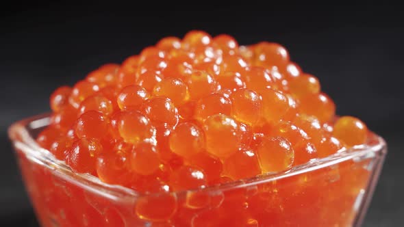 A Serving of Selected Red Caviar Rotates on a Black Background in a Transparent Glass Bowl