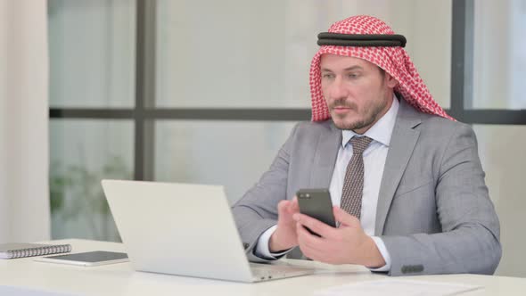 Middle Aged Arab Businessman using Smartphone while using Laptop in Office