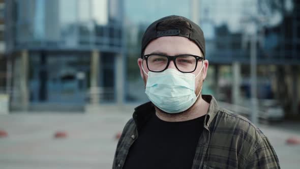 Portrait of Young Man in Glasses, Cap and Medical Mask Outdoors in the City