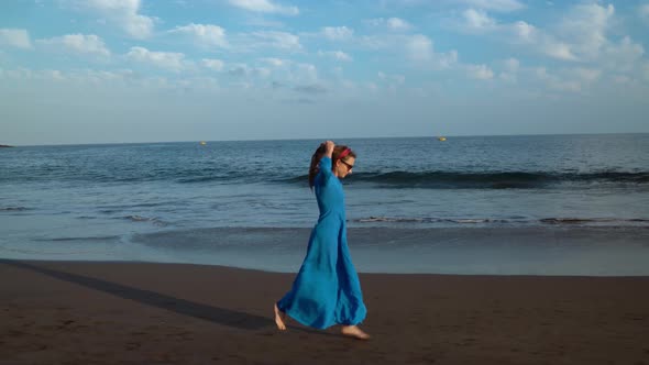 Woman in Sunglasses and Blue Dress Running Along a Black Volcanic Beach