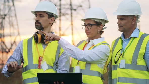 Engineers in Uniform Work with Laptop, Standing on Field with Power Lines.