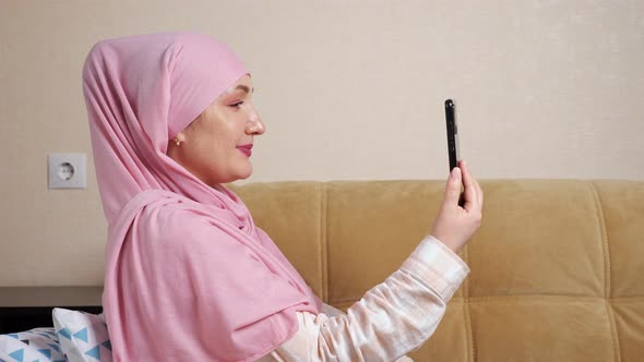 Young Muslim Woman in Pink Hijab Speaks By Video Call on the Phone While Sitting on the Couch