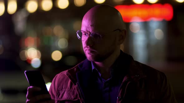 A Bald Solid Guy in a Brown Jacket Uses a Mobile Phone on the Street at Night