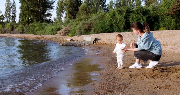 Mother teaching baby to throw stones in water sitting by the lake shore