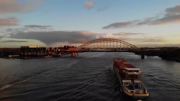 Freight Ship With Shipping Containers Sailing At Noord River With Arch Bridge In Background At Sunse