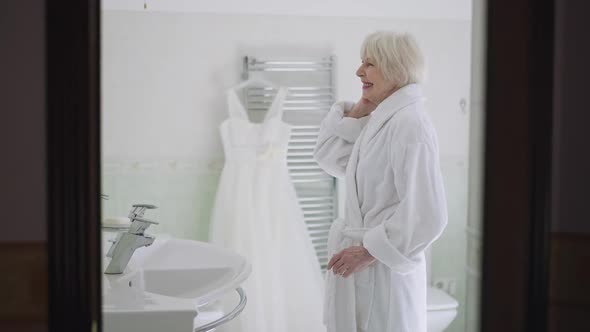 Happy Senior Caucasian Bride in Bathrobe Standing in Bathroom Admiring Reflection in Mirror and