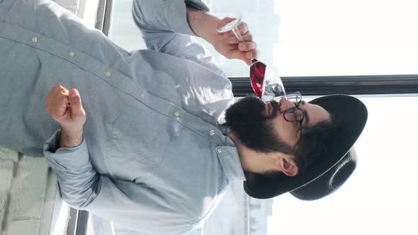 Pleased Bearded Man Drinking Red Wine and Admiring View Form Window at Home